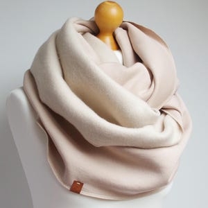 Pode incluir: Um cachecol infinito de tons neutros, creme e castanho claro, drapeado sobre um manequim. O cachecol é feito de um material espesso e quente, perfeito para o clima frio. Uma pequena etiqueta castanha é costurada na borda.