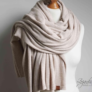 May include: A beige scarf draped over a mannequin. The scarf is made of a soft, knitted material and is wrapped around the mannequin's neck and shoulders. The scarf has a rectangular shape and a small, brown leather tag. The scarf is a neutral color.