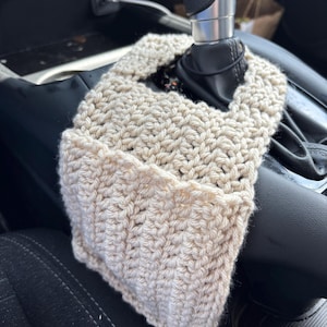 May include: A beige, hand-crocheted fabric cover designed to fit around a car gear shift lever. The accessory features a textured, ribbed stitch pattern and is constructed with an opening at the top to accommodate the gear shifter handle.