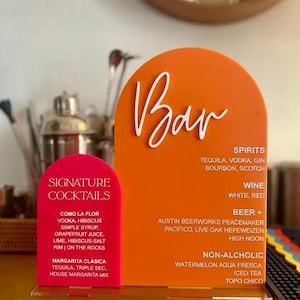 Może przedstawiać: Dwa akrylowe szyldy barowe. Jeden jest w kolorze jaskrawo pomarańczowym ze słowem "Bar" w białym skrypcie. Drugi to mniejszy, różowy znak z napisem "Signature Cocktails". Znaki wymieniają opcje napojów, w tym alkohole, wino, piwo i napoje bezalkoholowe.
