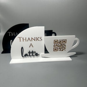 May include: A white and gold laser-cut sign with the words "THANKS a latte". The sign features a coffee cup with a QR code. A black sign with the word "THANKS" is visible in the background.
