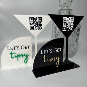 Könnte beinhalten: Martini-Glas-förmige Schilder aus weißem und schwarzem Acryl mit QR-Codes und dem Schriftzug "LET'S GET tipsy". Ein Schild hat goldene Buchstaben, das andere grüne. Im Hintergrund ist eine klare Glas-Karaffe zu sehen.