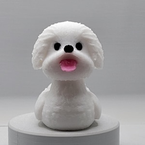 Może przedstawiać: Biała ceramiczna figurka psa z czarnymi oczami i wystającym różowym językiem. Pies siedzi na białej podstawie.