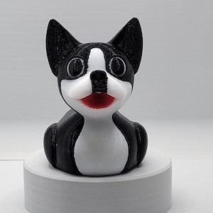 May include: A black and white 3D printed figurine of a Boston Terrier dog. The dog is sitting with its head tilted up and its mouth slightly open, revealing a red tongue. The figurine has a white patch on its chest and white markings around its eyes.