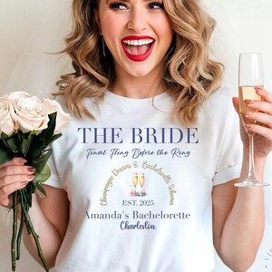 May include: A white t-shirt with the text "THE BRIDE" and "Final Fling Before the Ring". The shirt also features a graphic and text for a bachelorette party in Charleston, EST. 2025. The person is wearing a tiara and holding a bouquet of roses and a champagne flute.