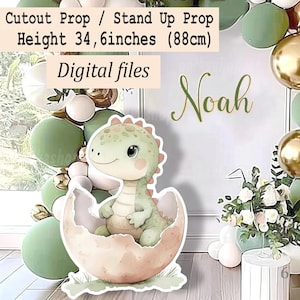 May include: A cutout prop of a baby dinosaur hatching from an egg, with the text "Cutout Prop / Stand Up Prop Height 34,6 inches (88cm) Digital files Noah". The dinosaur is light green with pink accents. The background includes balloons and floral arrangements.