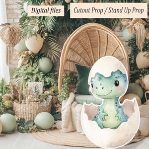 Pode incluir: Ilustração em aquarela de um bebê dinossauro saindo de um ovo, em tons de verde e azul. A imagem é um recorte, com o texto "Arquivos digitais" e "Adereço recortado / Adereço de pé". O fundo inclui uma cadeira de vime, vegetação e balões.