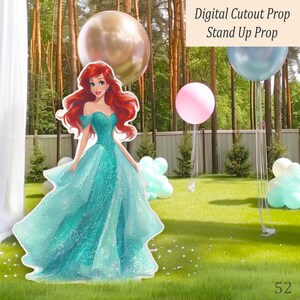 A cardboard cutout of a Mermaid princess in a teal gown with red hair, set on a lawn with balloons. The image includes the text "Digital Cutout Prop Stand Up Prop". The scene is set outdoors with trees in the background.