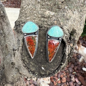 Op de afbeelding: Een paar handgemaakte oorbellen met een turquoise bovenkant en een rood-oranje patroonsteen. De oorbellen hebben een zilveren metalen rand en zijn geplaatst tegen een natuurlijke houten achtergrond.