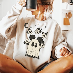 May include: Cream-colored sweatshirt featuring a Halloween-themed graphic. The design includes a ghost with Mickey Mouse ears, surrounded by skulls and a crescent moon. The sweatshirt is paired with denim shorts and fuzzy slippers.