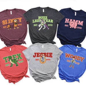 Op de afbeelding: Zes kleurrijke t-shirts met personages uit een populaire animatiefilm. Elk shirt heeft de naam van een personage en het oprichtingsjaar. De shirts zijn in verschillende kleuren, waaronder bordeauxrood, grijs, marineblauw, rood, grijs en blauw.