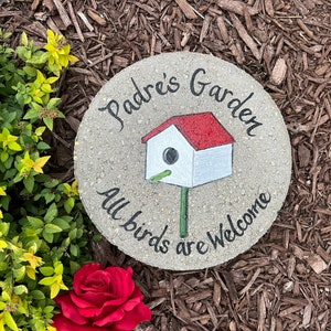 May include: A round concrete garden stone with the words "Padre's Garden" and "All birds are welcome" painted in black. A white birdhouse with a red roof is painted in the centre of the stone.