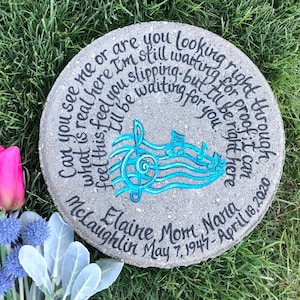 May include: A gray concrete memorial stone with a blue musical note design and the inscription "Can you see me or are you looking right through? What is real here I'm still waiting for you. Feel this feel you slipping - but I'll be proof it can be right here.  Elaine Mom Nara McLaughlin May 7, 1947 - April 18, 2020"