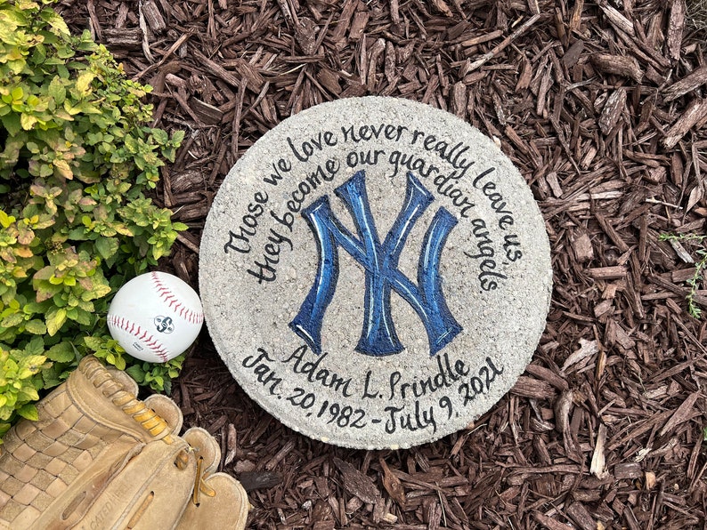Puede incluir: Un memorial de piedra gris con un logotipo de los New York Yankees azul y blanco pintado en &eacute;l. La piedra tiene el texto "Those we love never really leave us they become our guardian angels" y "Adam L. Prindle Jan. 20, 1982 - July 9, 2021".