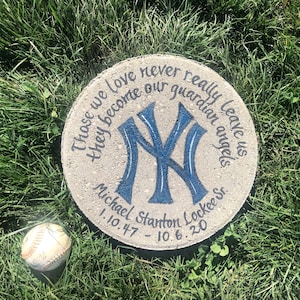 Puede incluir: Una piedra conmemorativa redonda de hormig&oacute;n con un logotipo de los New York Yankees azul y blanco pintado en ella. La piedra tambi&eacute;n tiene el texto "Those we love never really leave us they become our guardian angels" y "Michael Stanton Lockee Sr. 1.10.47 - 10.6.20".