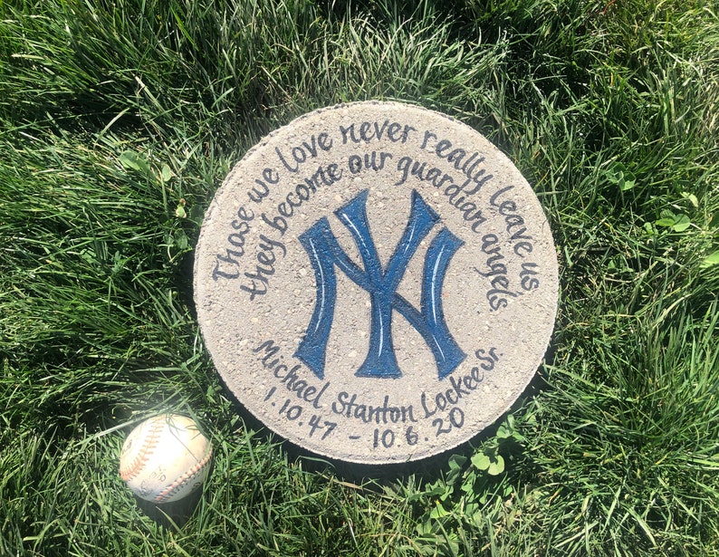Puede incluir: Una piedra conmemorativa redonda de hormig&oacute;n con un logotipo de los New York Yankees azul y blanco pintado en ella. La piedra tambi&eacute;n tiene el texto "Those we love never really leave us they become our guardian angels" y "Michael Stanton Lockee Sr. 1.10.47 - 10.6.20".