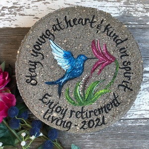 May include: A round concrete stepping stone with a painted hummingbird, flowers, and the text "Stay young at heart & kind in spirit. Enjoy retirement living - 2021".