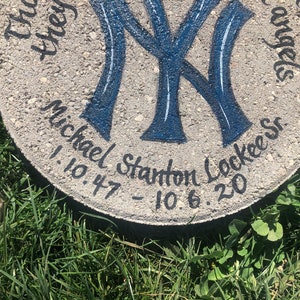 Puede incluir: Un adoqu&iacute;n de concreto gris con un logotipo de los New York Yankees azul y blanco pintado en &eacute;l. El texto "Michael Stanton Lockee" y las fechas "1.10.47 - 10.6.20" est&aacute;n escritas en pintura negra alrededor del logotipo.