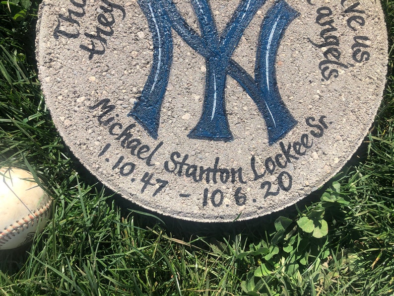 Puede incluir: Un adoqu&iacute;n de concreto gris con un logotipo de los New York Yankees azul y blanco pintado en &eacute;l. El texto "Michael Stanton Lockee" y las fechas "1.10.47 - 10.6.20" est&aacute;n escritas en pintura negra alrededor del logotipo.
