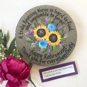 Puede incluir: Una piedra redonda gris con un diseño pintado de girasoles y flores moradas. La piedra tiene el texto "A truly amazing Nurse is hard to find and impossible to forget. Happy Retirement. Thank you for everything Shelly."