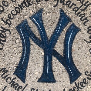 Puede incluir: Un logotipo azul y blanco de los New York Yankees pintado en una superficie de hormig&oacute;n gris. Las palabras "they become our guardian angels" y "michael/sharon/yankees" est&aacute;n escritas alrededor del logotipo.