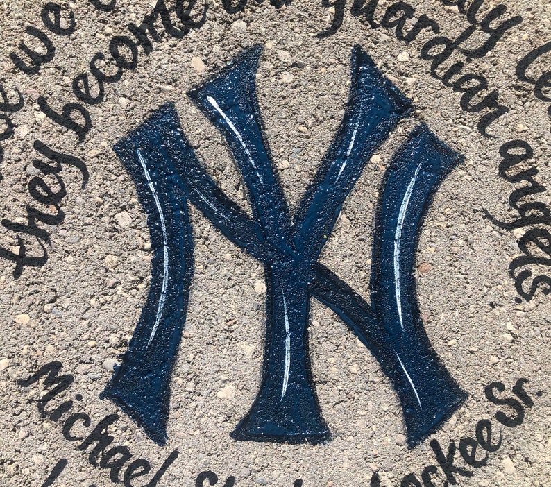 Puede incluir: Un logotipo azul y blanco de los New York Yankees pintado en una superficie de hormig&oacute;n gris. Las palabras "they become our guardian angels" y "michael/sharon/yankees" est&aacute;n escritas alrededor del logotipo.