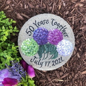 Puede incluir: Una piedra redonda gris con las palabras "50 Years Together" y "July 17, 2021" pintadas en ella. La piedra está decorada con hortensias pintadas de color morado, azul y verde.