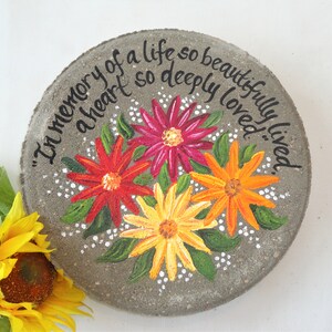 Puede incluir: Una piedra conmemorativa de hormigón pintada con el texto "In memory of a life so beautifully lived, a heart so deeply loved." La piedra está decorada con tres flores pintadas en tonos rojos, naranjas y amarillos.
