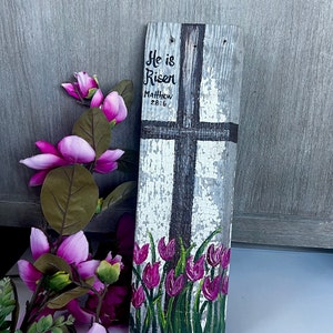 Puede incluir: Un letrero de madera con una cruz pintada y las palabras "He is Risen" y "Matthew 28:6". El letrero está decorado con tulipanes morados pintados.