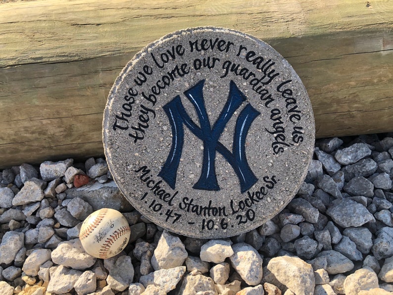 Puede incluir: Una piedra conmemorativa de hormig&oacute;n gris con un logotipo de los New York Yankees azul y blanco pintado en ella. La piedra tiene el texto "Those we love never really leave us they become our guardian angels" y "Michael Stanton Lockee Sr. 1.10.47 - 10.6.20" escrito en ella.