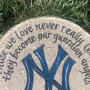 Puede incluir: Un adoqu&iacute;n de hormig&oacute;n gris con una inscripci&oacute;n pintada en negro que dice "Those we love never really leave us they become our guardian angels." La piedra tambi&eacute;n presenta un logotipo de los New York Yankees pintado en azul y blanco.