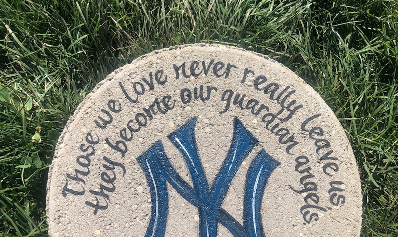 Puede incluir: Un adoqu&iacute;n de hormig&oacute;n gris con una inscripci&oacute;n pintada en negro que dice "Those we love never really leave us they become our guardian angels." La piedra tambi&eacute;n presenta un logotipo de los New York Yankees pintado en azul y blanco.