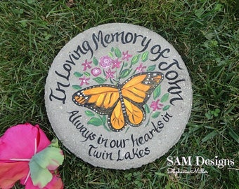 May include: A gray stone with the words "In Loving Memory of John Always in our hearts in Twin Lakes" painted on it. A painted butterfly with orange and black wings is in the center of the stone. The stone is surrounded by green grass.