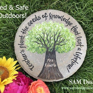 May include: A round, grey stone painted with a green tree and the text "Teacher's plant the seeds of knowledge that last a lifetime". The text "Mrs. Roberts" is written below the tree.