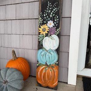 Peut inclure: Un panneau en bois rectangulaire et haut avec un fond noir, illustré d'une pile verticale de trois citrouilles blanche, bleu clair et orange. Au-dessus des citrouilles sont peints des tournesols jaunes, des fleurs violettes et des tiges vertes.