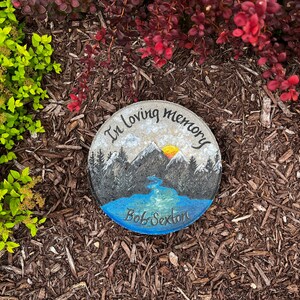 May include: A painted memorial stone with a mountain scene and a river flowing through it. The stone reads "In loving memory Bob Sexton".