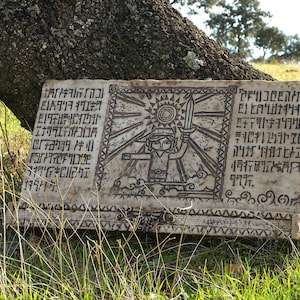 Puede incluir: Placa rectangular de piedra blanquecina con una ilustración negra de un personaje sosteniendo una espada, sobre un fondo de rayos solares. La placa, con texto antiguo, descansa sobre hierba cerca de un tronco de árbol. Tema de fantasía.