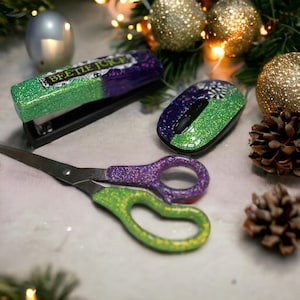 May include: A set of green and purple glitter office supplies, including a stapler, a computer mouse, and a pair of scissors. The stapler has the words "Beetle Juice" written on it.