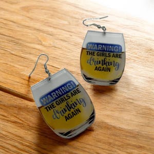 May include: A pair of novelty earrings shaped like beer glasses. Each earring has a silver hook and features the text "WARNING! THE GIRLS ARE drinking AGAIN" in blue and cursive font over a yellow and clear background.