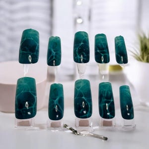 May include: Set of ten artificial nails with a teal and white marbled design. Each nail is displayed on a clear stand, showcasing the intricate pattern. A silver tool rests near the base of the display, suggesting nail art application.