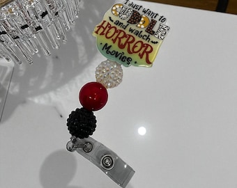May include: A retractable badge reel with the text "I just want to cuddle and watch HORROR Movies." The reel features a yellow and green design, with a red, black, and white beaded accent. The reel has a clear plastic clip.