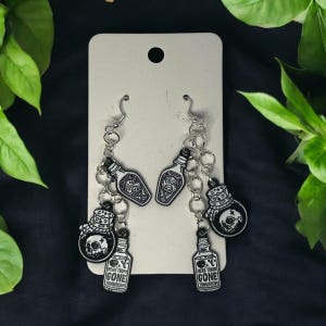 May include: A pair of silver chain earrings with black and white charms. The charms are shaped like bottles and jars with the text "Here Today Gone Tomorrow" and a skull and crossbones.