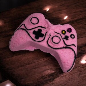 May include: A pink bath bomb shaped like a video game controller. The controller has black line details and a black cross button. Other buttons are colored yellow, purple, and pink. The bath bomb is on a wooden surface.