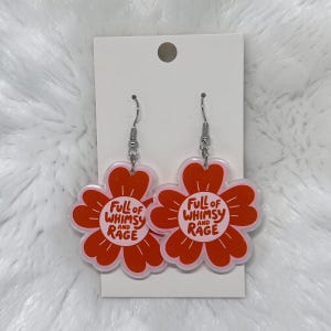 May include: A pair of dangle earrings featuring orange flower-shaped charms with a light pink center. The text "FULL OF WHIMSY AND RAGE" is printed in orange on the center of each flower. The earrings are attached to a white rectangular display card.