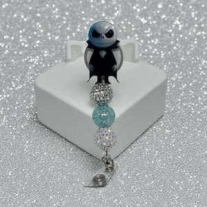May include: A retractable badge reel featuring a Jack Skellington character design. The reel has a black and white character image, with a series of decorative beads in silver and blue. The reel is attached to a clear plastic clip.