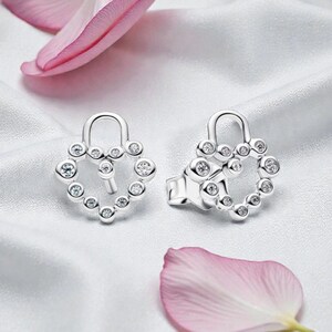 May include: A pair of silver-colored stud earrings. Each earring features a heart-shaped design with a keyhole and is adorned with small, clear gemstones. The earrings are displayed on a white, silky surface, with pink flower petals in the background.
