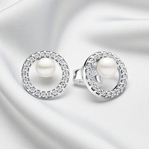 Peut inclure: Une paire de boucles d'oreilles à clous argentées. Chaque boucle d'oreille présente un centre de perle blanche entouré d'un halo de petits cristaux transparents. Les boucles d'oreilles sont posées sur un fond blanc et soyeux.