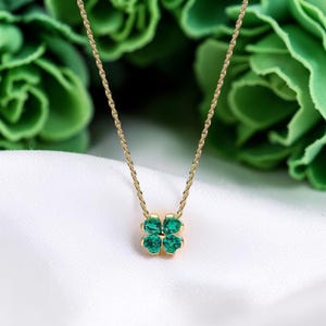 Puede incluir: Un collar dorado con un colgante de trébol de cuatro hojas. El trébol está hecho de cuatro piedras preciosas de color verde esmeralda. El collar se muestra sobre una superficie de satén blanco con un fondo verde borroso.
