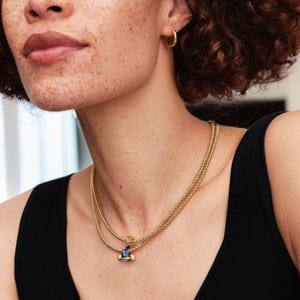 May include: A gold-toned necklace with a double chain design. The necklace features a pendant with blue and gold accents. The person in the image is wearing a black top and gold hoop earrings.