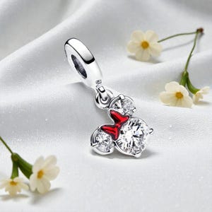 May include: A silver charm with a Minnie Mouse design. The charm features three clear, sparkling cubic zirconia stones and a red enamel bow. The charm is suspended from a silver loop, and is set against a white, silky background.